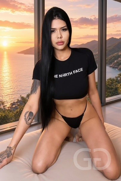 Angel, busty Latin escort, in a black crop tee with a sunset sea view behind