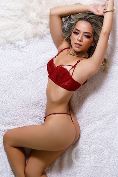 Lola, high class Latin escort London, lying on a white quilt in red lace lingerie