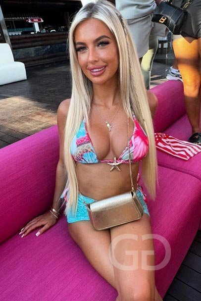 Alana, English party girl escort, on a pink sofa in a tropical bikini